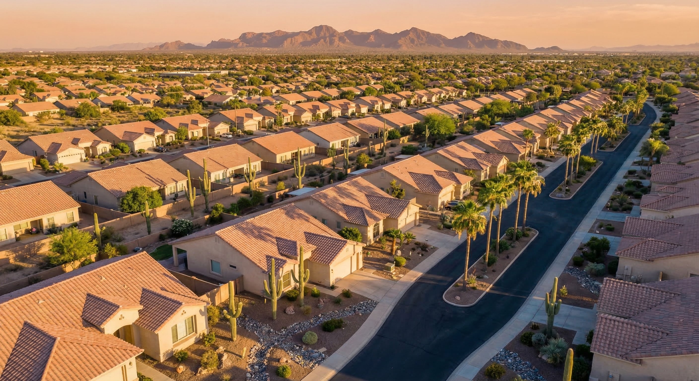 Aerial drone view of sprawling Phoenix suburban neighborhoods with rows of stucco ranch homes, terracotta tile roofs, desert landscaping, and mountain backdrop at golden hour