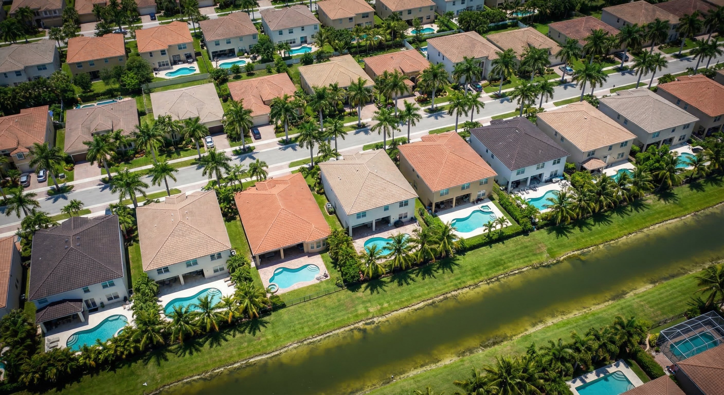 Aerial drone photograph from 400 feet over a South Florida neighborhood showing rows of stucco homes with flat roofs and blue backyard swimming pools, palm trees lining the streets, a canal visible running through the frame