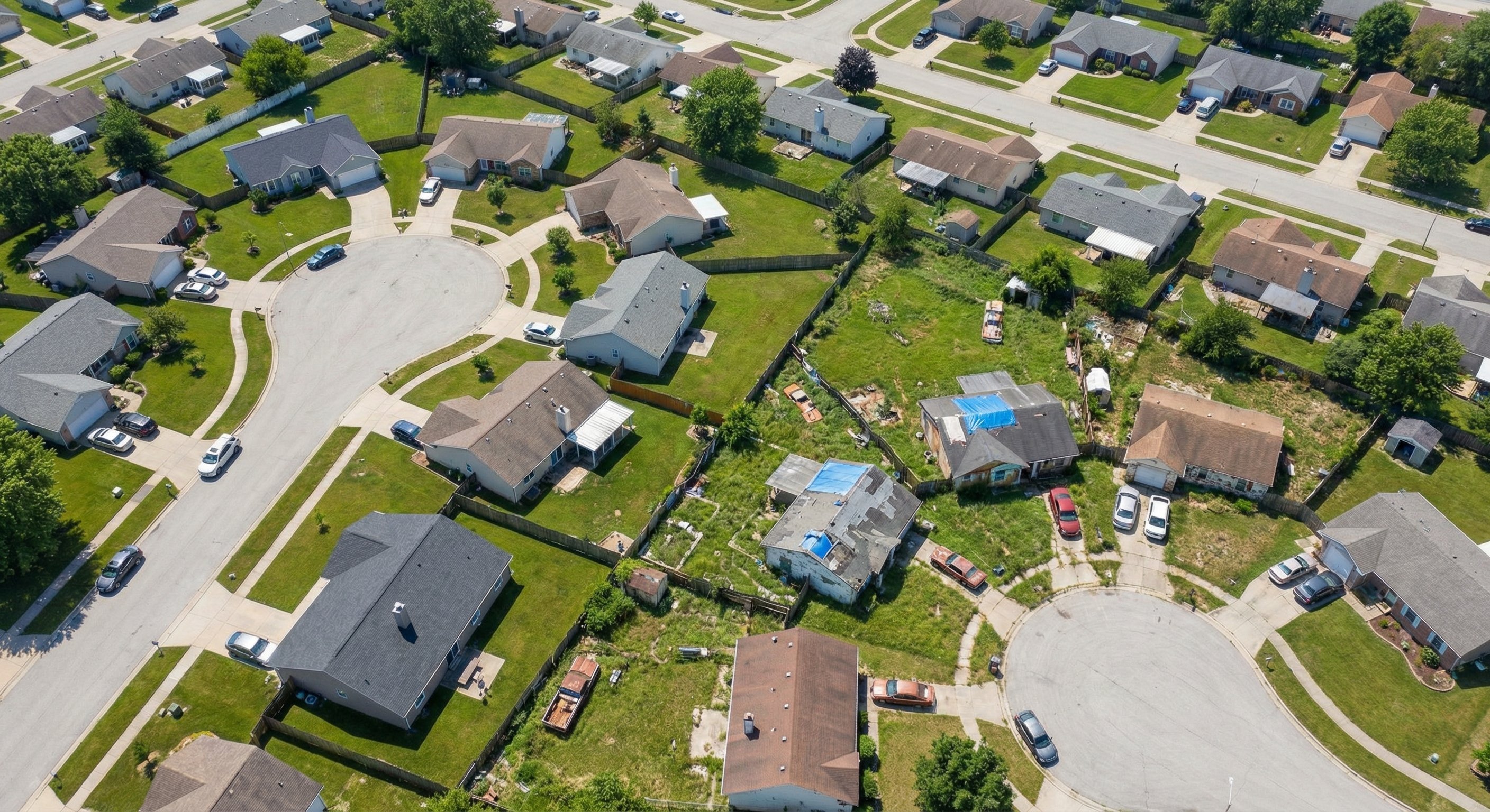 Aerial drone photograph of a residential neighborhood showing variation in property conditions, some well-maintained homes next to clearly distressed properties with overgrown lots
