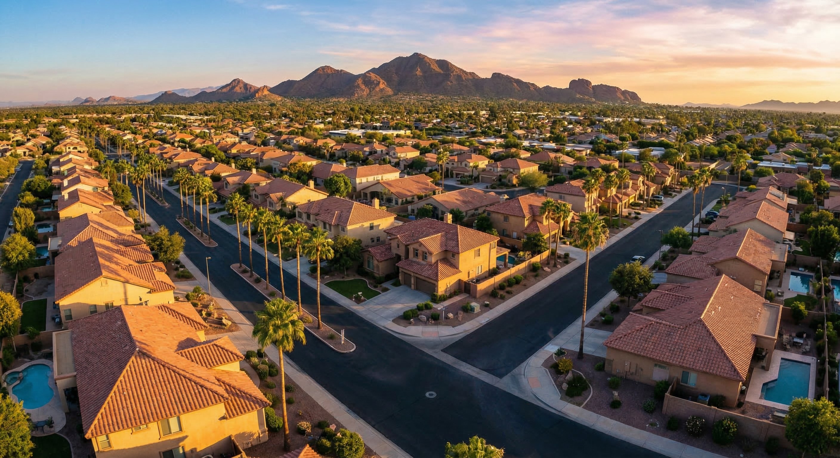 Aerial view of a Phoenix-area residential neighborhood in late afternoon light, terracotta rooftops and stucco homes visible, desert mountains in the background, palm trees lining wide streets, DJI Mavic 3 wide angle photography, real estate aerial