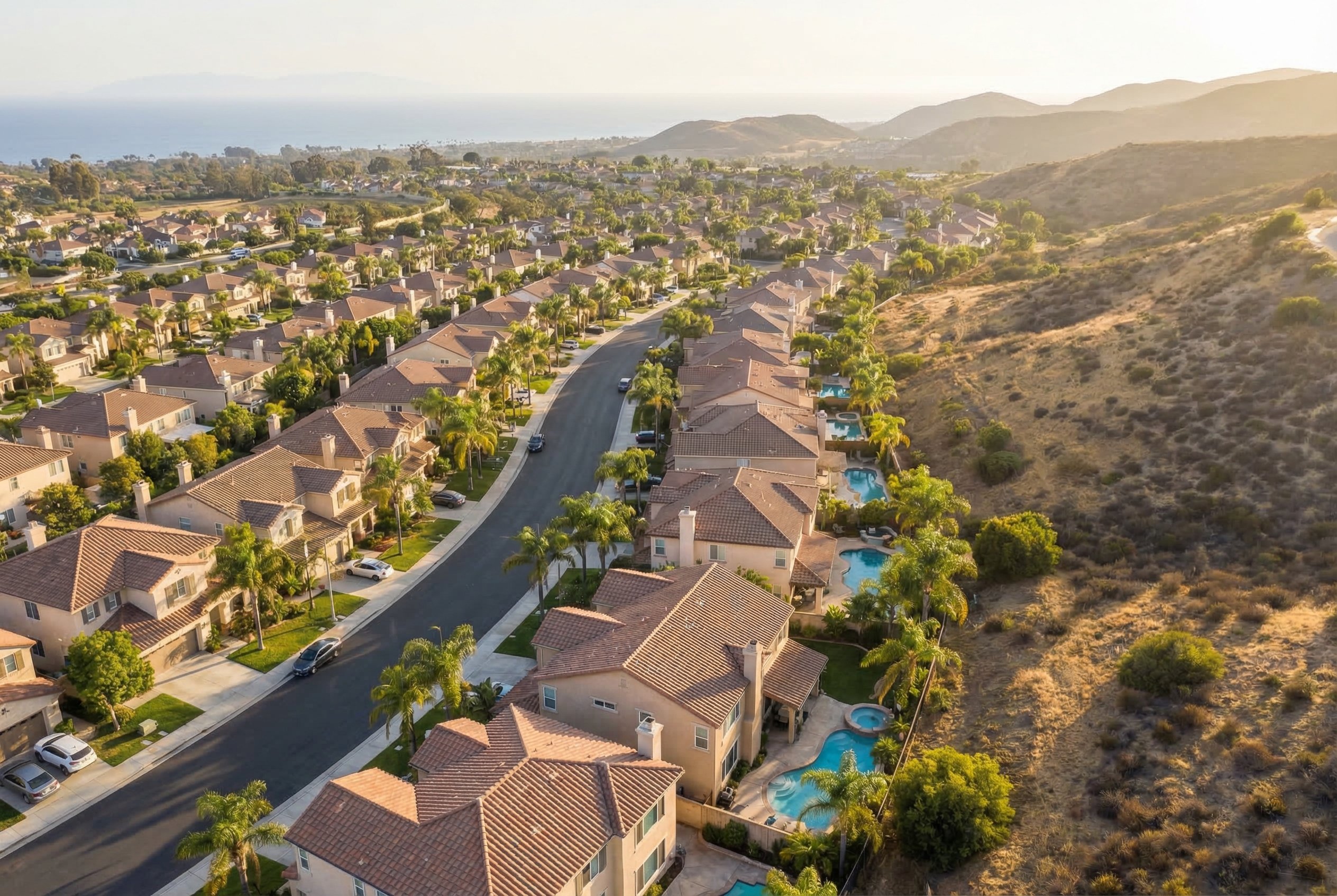 Aerial drone photograph from 800 feet looking over a dense Southern California residential neighborhood, grid of stucco homes with tile roofs visible, palm-lined streets and dry hills in background, warm afternoon light, DJI Mavic 3 wide angle, real estate aerial photography
