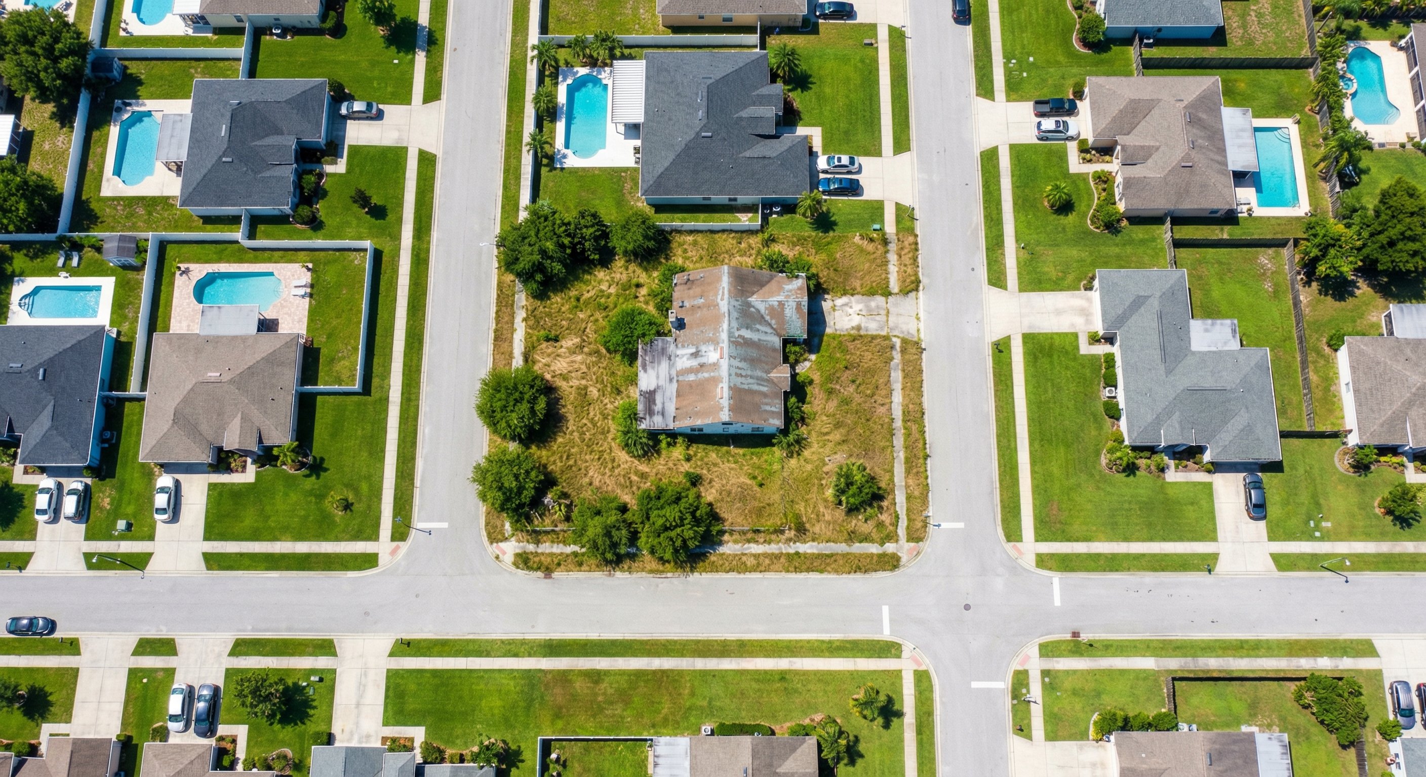Aerial view of a residential neighborhood showing one vacant overgrown property contrasting with well-maintained homes