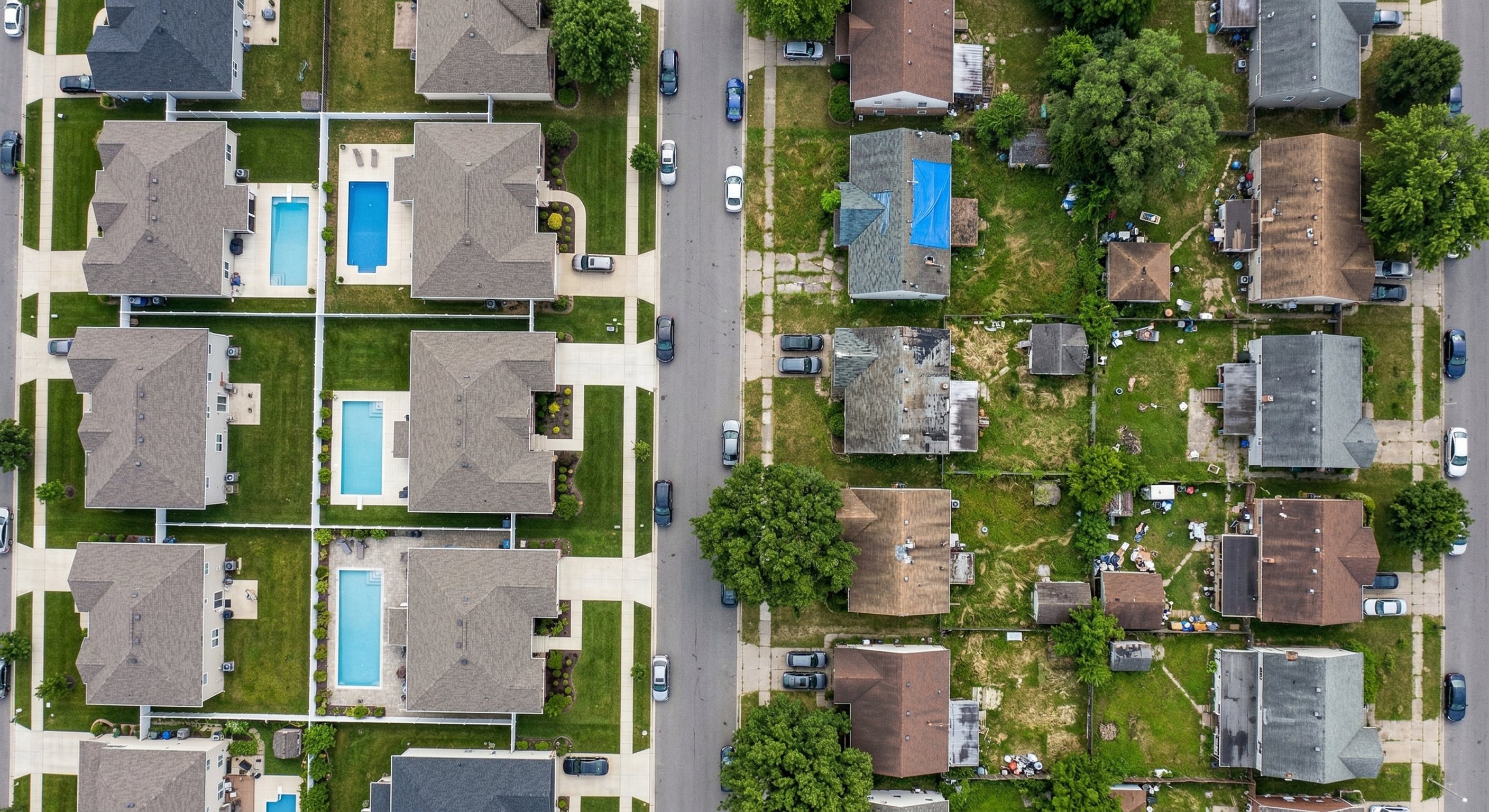An aerial photograph from 400 feet looking down at a suburban residential neighborhood with varied housing conditions. Some properties appear well-maintained, others show overgrown yards or aging roofs. Real estate aerial photography, midday light.