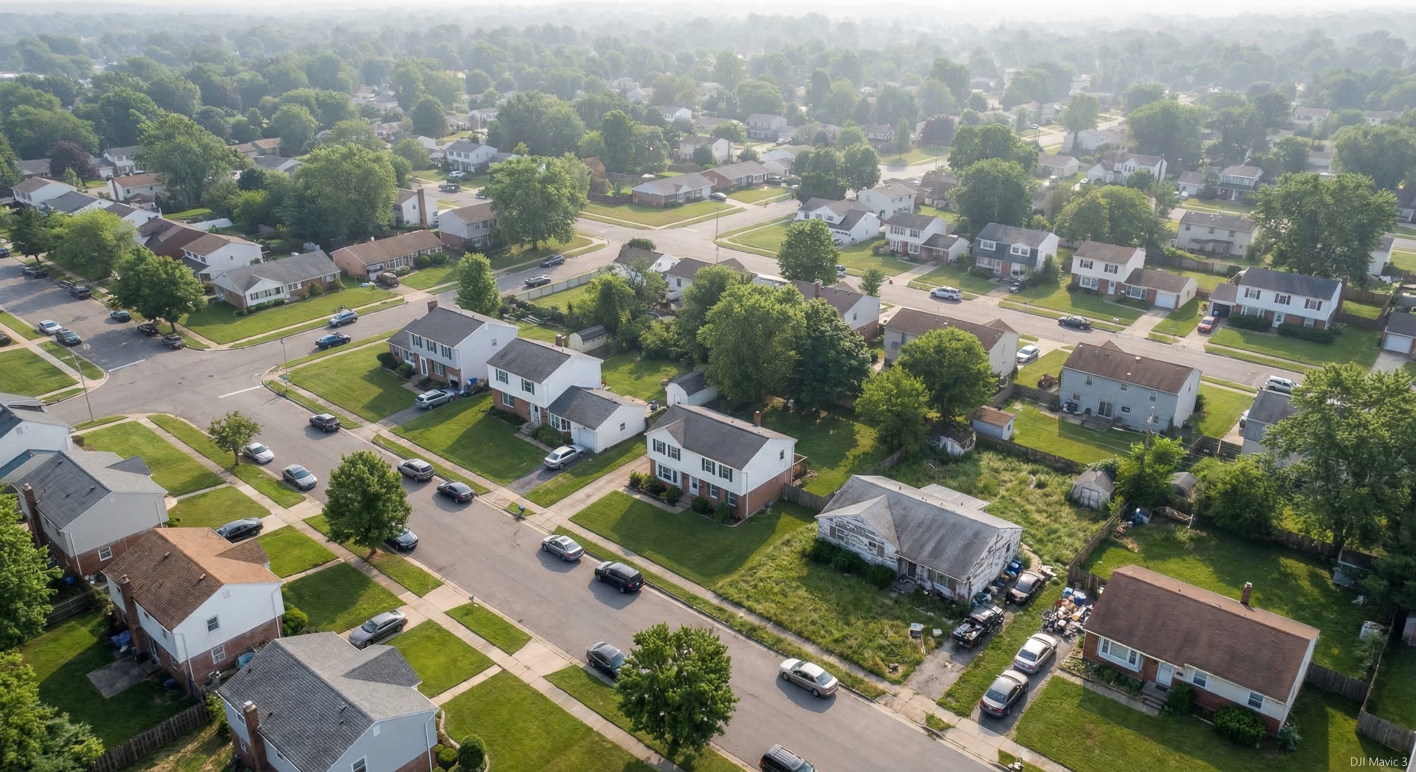 Aerial drone photograph showing a residential neighborhood with varied home styles, mix of well-maintained properties and homes with visible deferred maintenance, suburban setting with tree-lined streets
