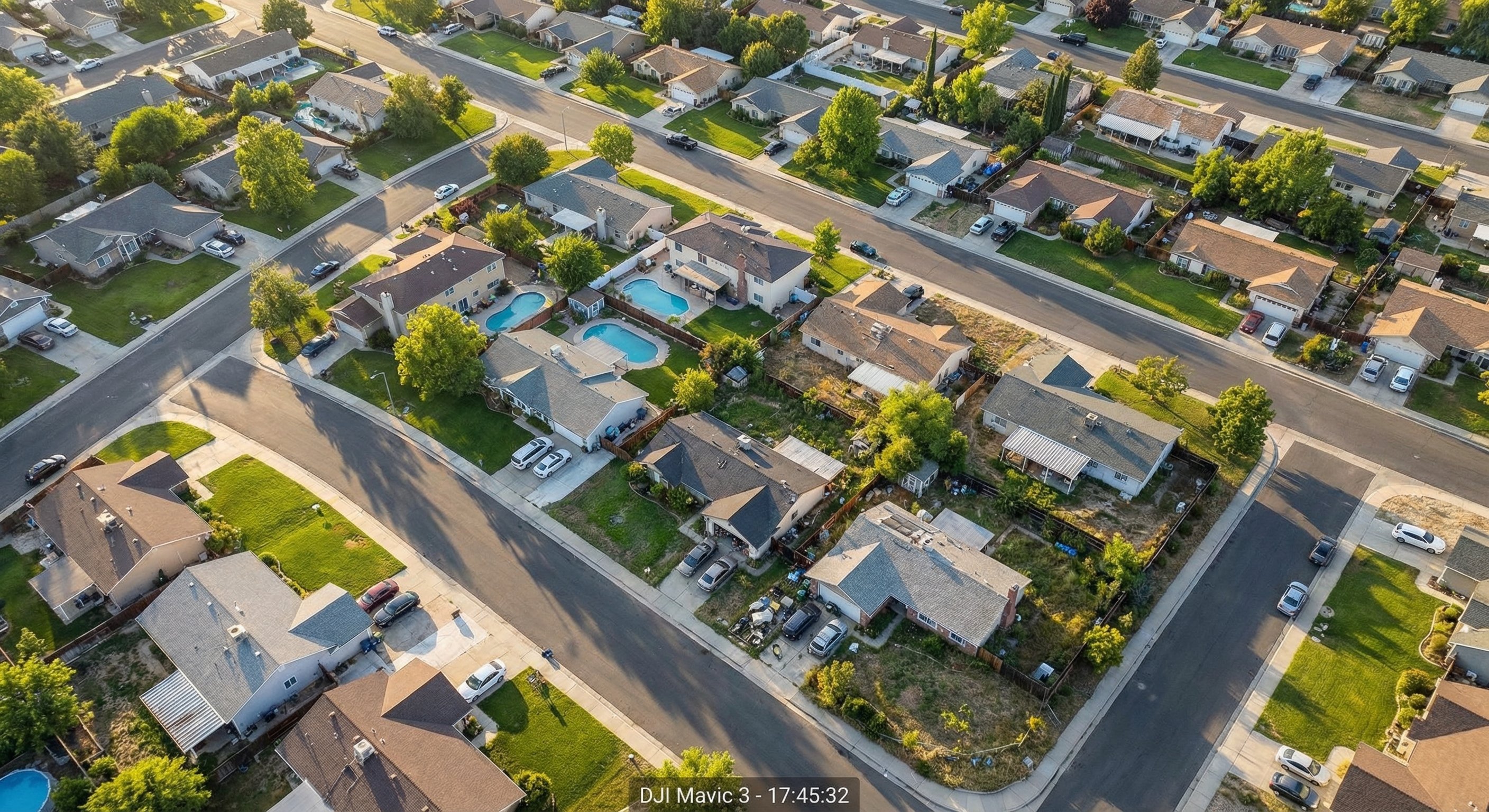 Aerial drone photograph from 200 feet showing a sprawling suburban neighborhood with varied rooflines and property conditions, late afternoon light casting long shadows across the streets