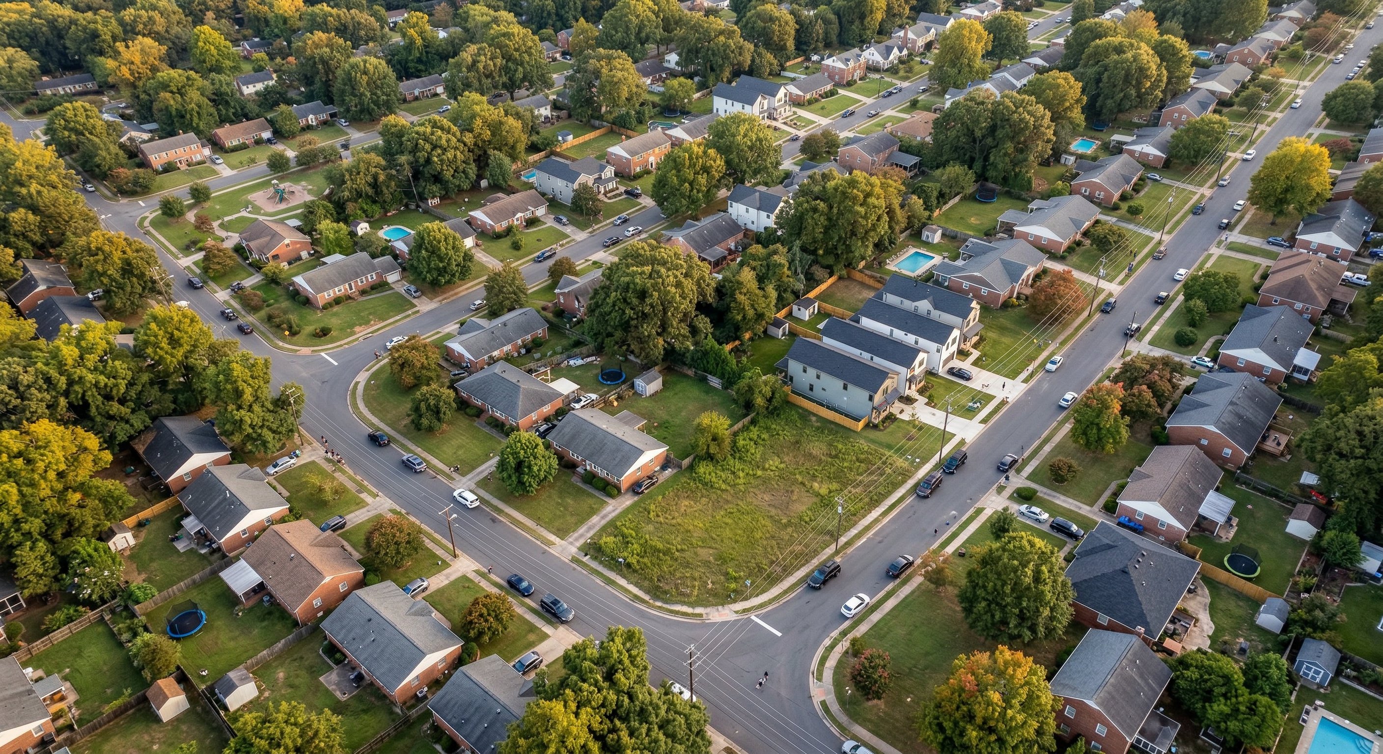 Aerial drone view looking down at a mixed residential neighborhood in the Charlotte-Mecklenburg metro area, suburban streets lined with trees, warm afternoon light, DJI Mavic 3 aerial photography