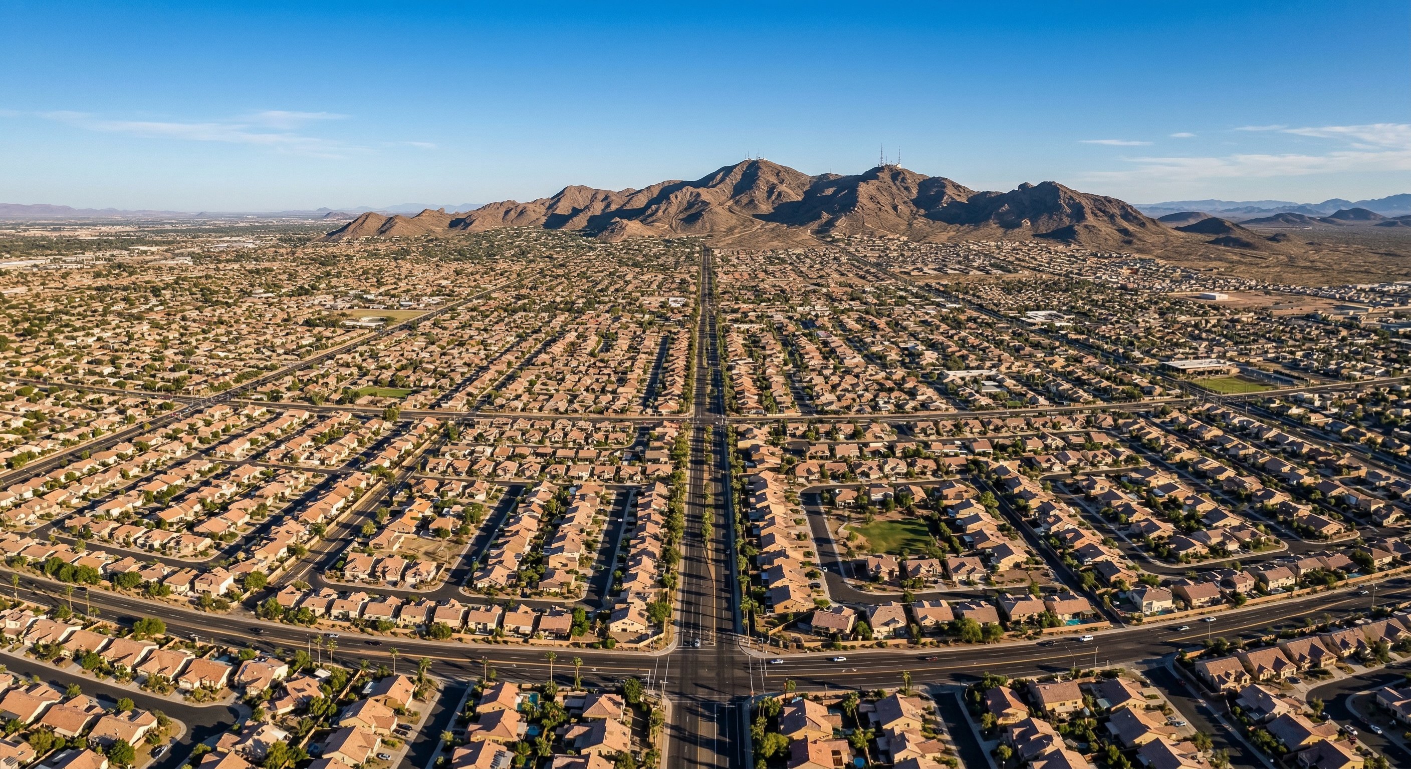 Stylized aerial drone photograph of the greater Phoenix metro area from 2,000 feet, looking south at a dense suburban grid of tan stucco subdivisions extending toward South Mountain, showing the vast scale of Maricopa County's housing stock in warm late-afternoon Arizona sunlight