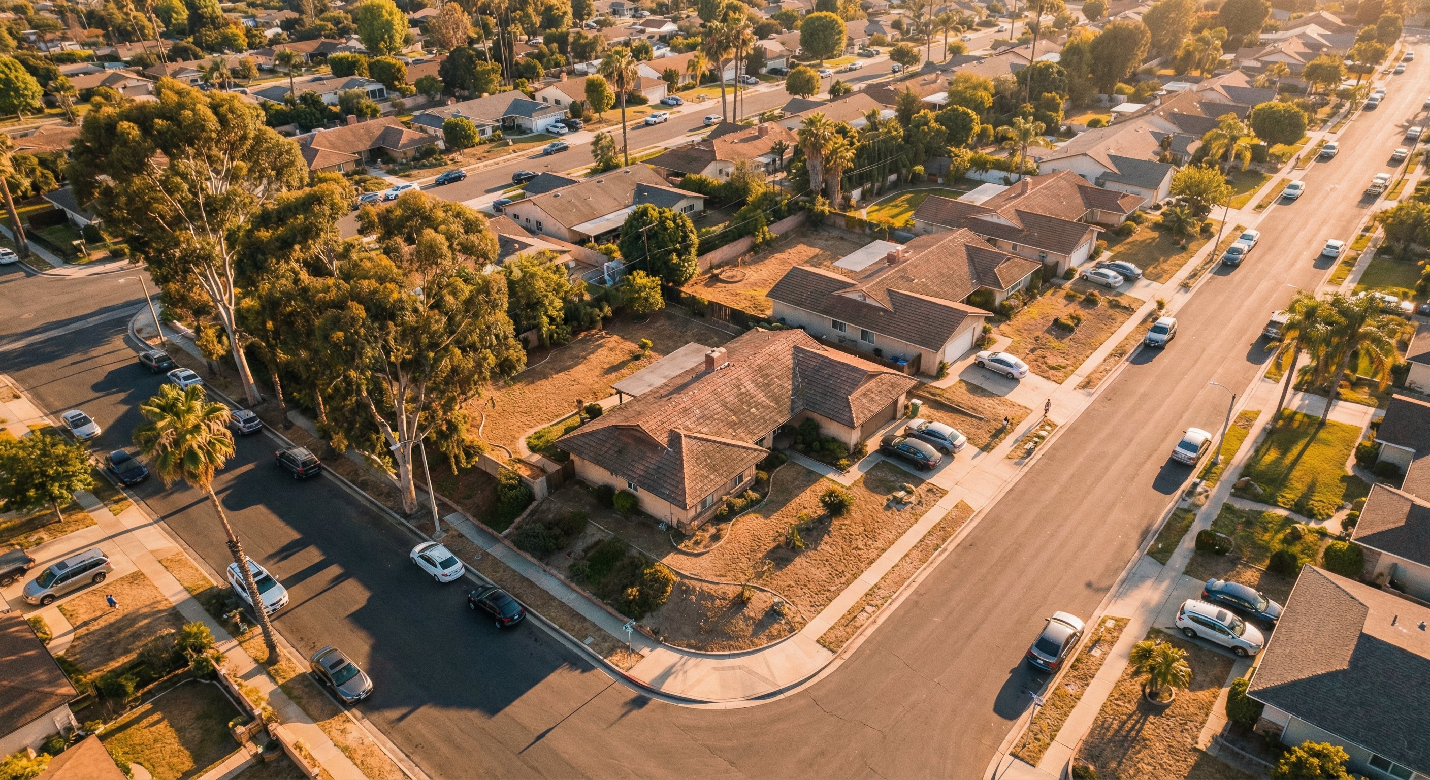 Aerial drone photograph of a California residential neighborhood at golden hour, sprawling single-story ranch homes with tile roofs, mature trees, long shadows from late afternoon sun. Real estate investment market overview, wide angle, rich warm light, documentary aerial photography.