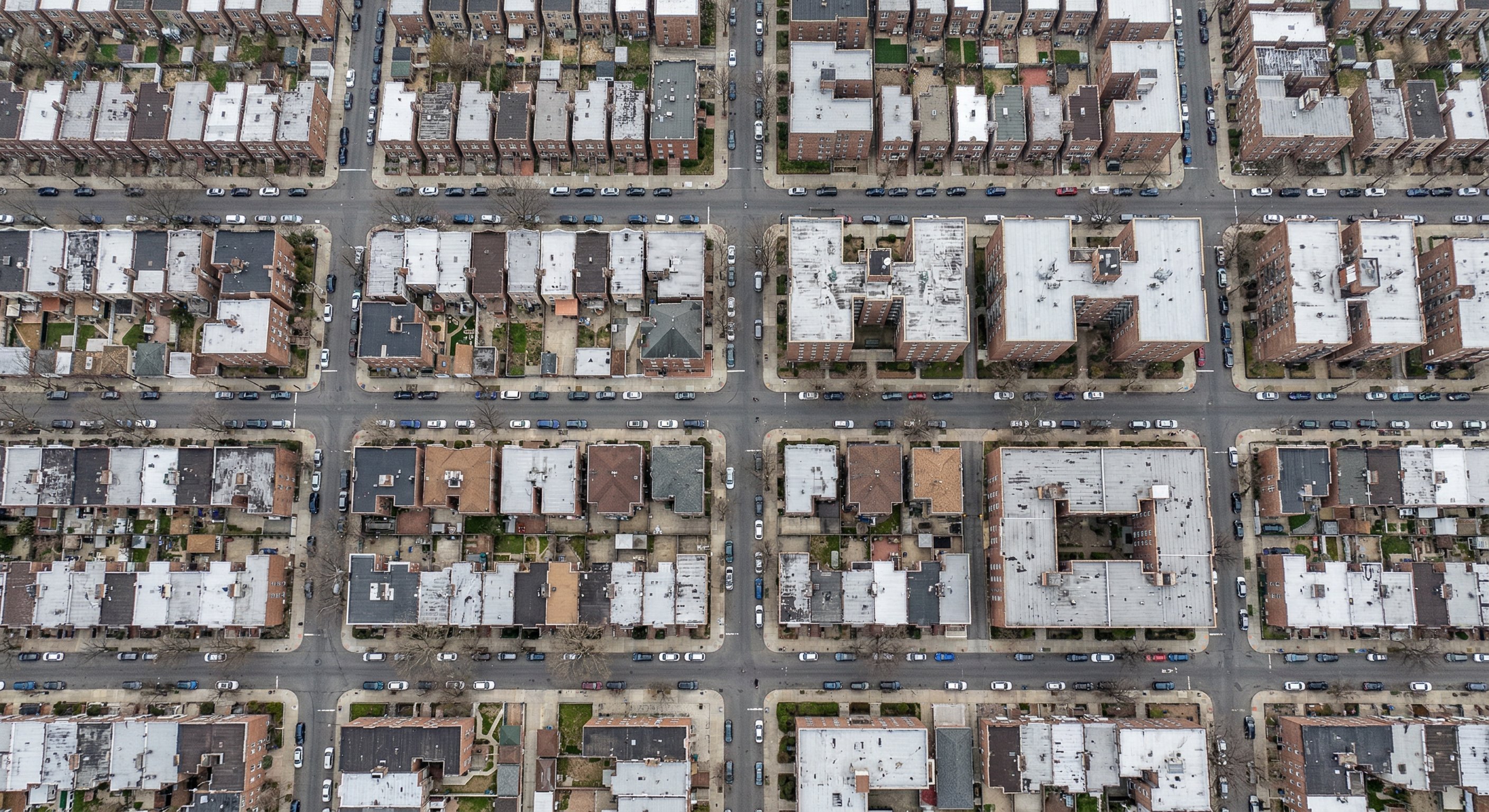Aerial drone photograph of outer borough New York neighborhood showing Queens street grid with brick rowhouses and apartment buildings, overcast early spring light, DJI Mavic 3 wide-angle. Real estate aerial photography.