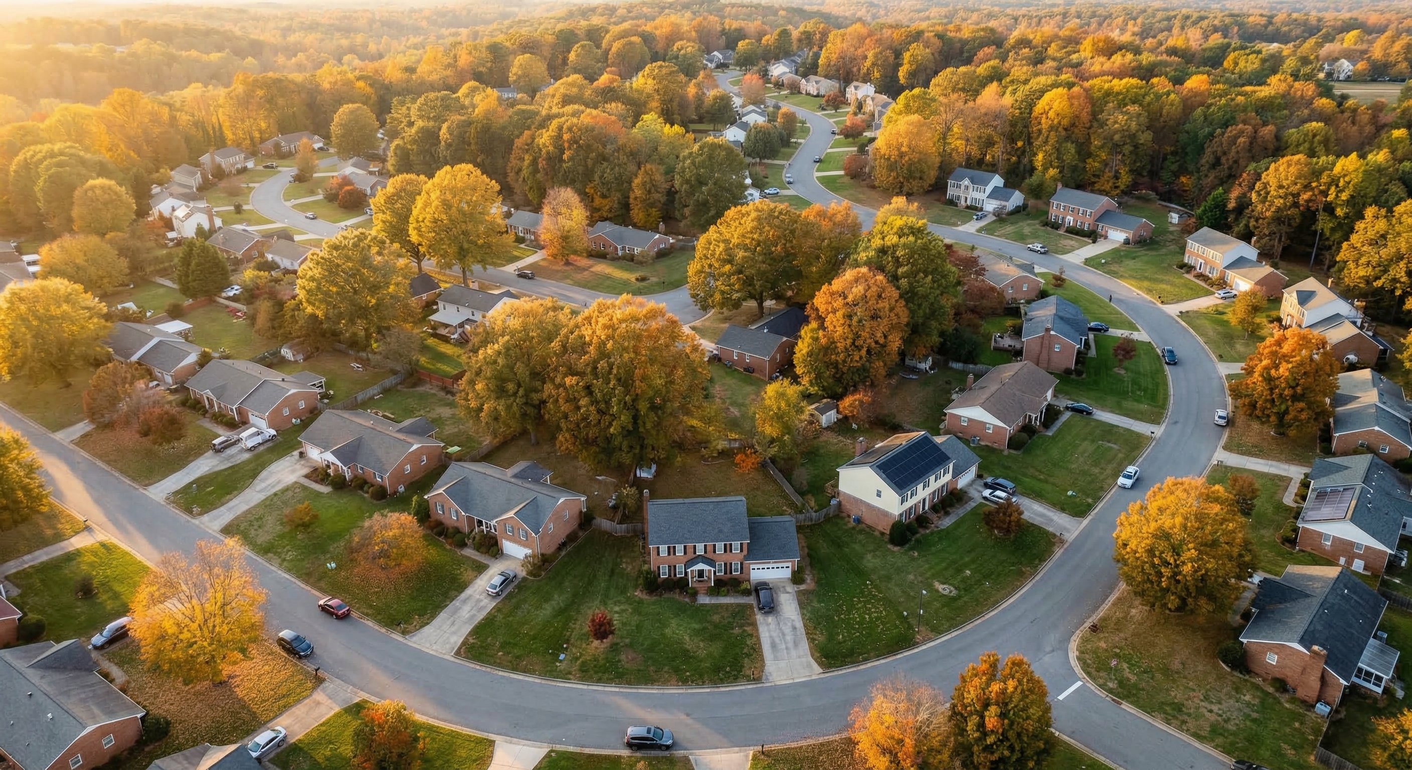 Aerial drone view of a North Carolina Piedmont subdivision with brick ranch homes, mature oak trees in fall color, and curving suburban streets