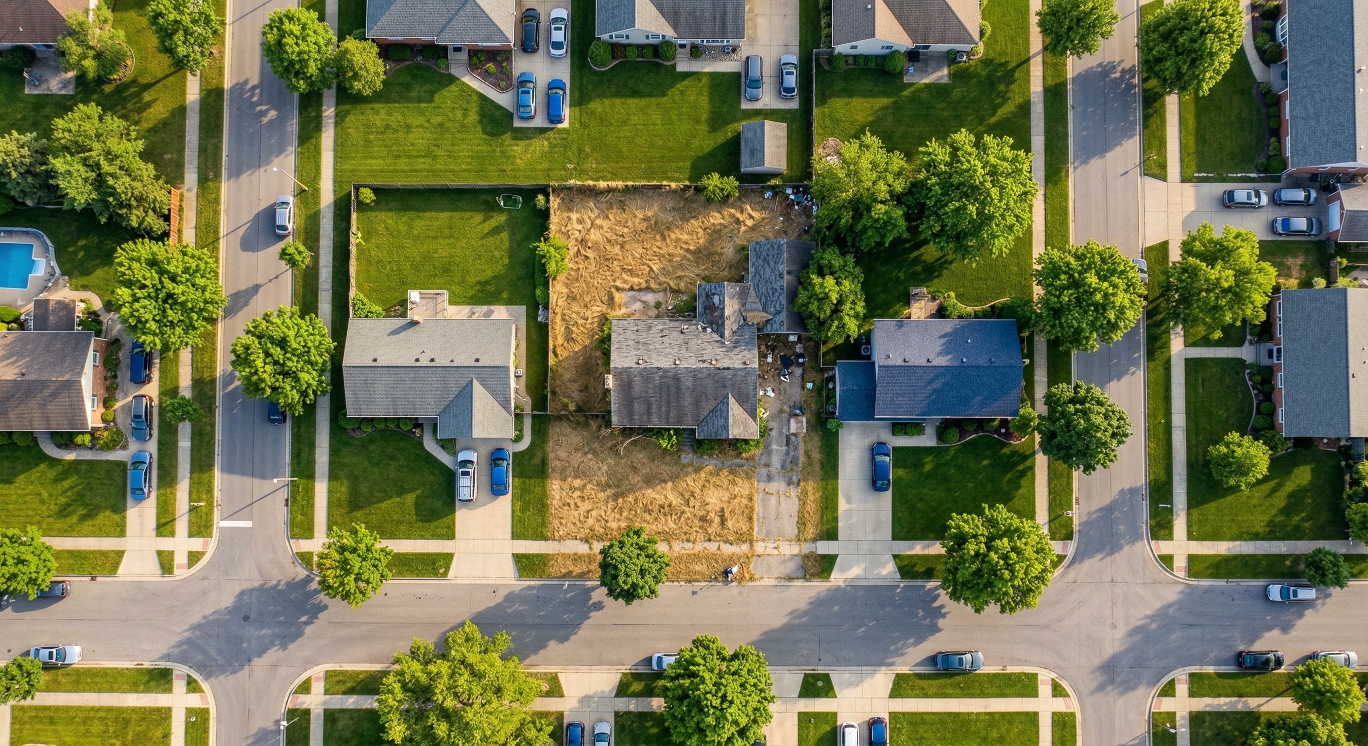 Aerial drone photograph from 400 feet showing a residential block in a mid-sized American city, where one visibly neglected property with an overgrown yard and weathered exterior stands out among well-maintained neighboring homes. Late afternoon golden hour light. Residential neighborhood context. DJI Mavic 3 wide angle. Real estate aerial photography.