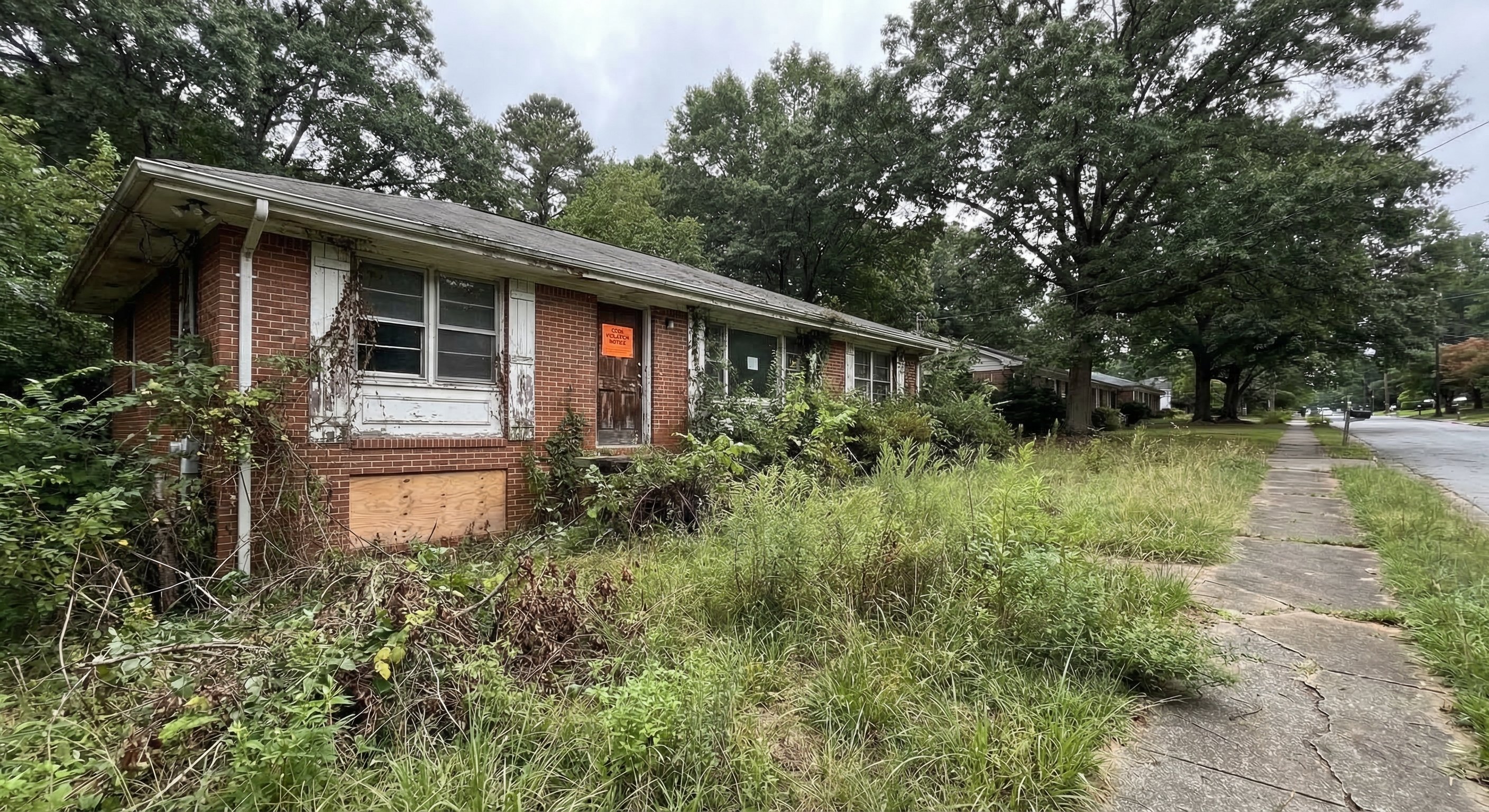 Vacant 1970s brick ranch home in suburban Atlanta with overgrown lawn, peeling trim, and code violation notice posted on front door