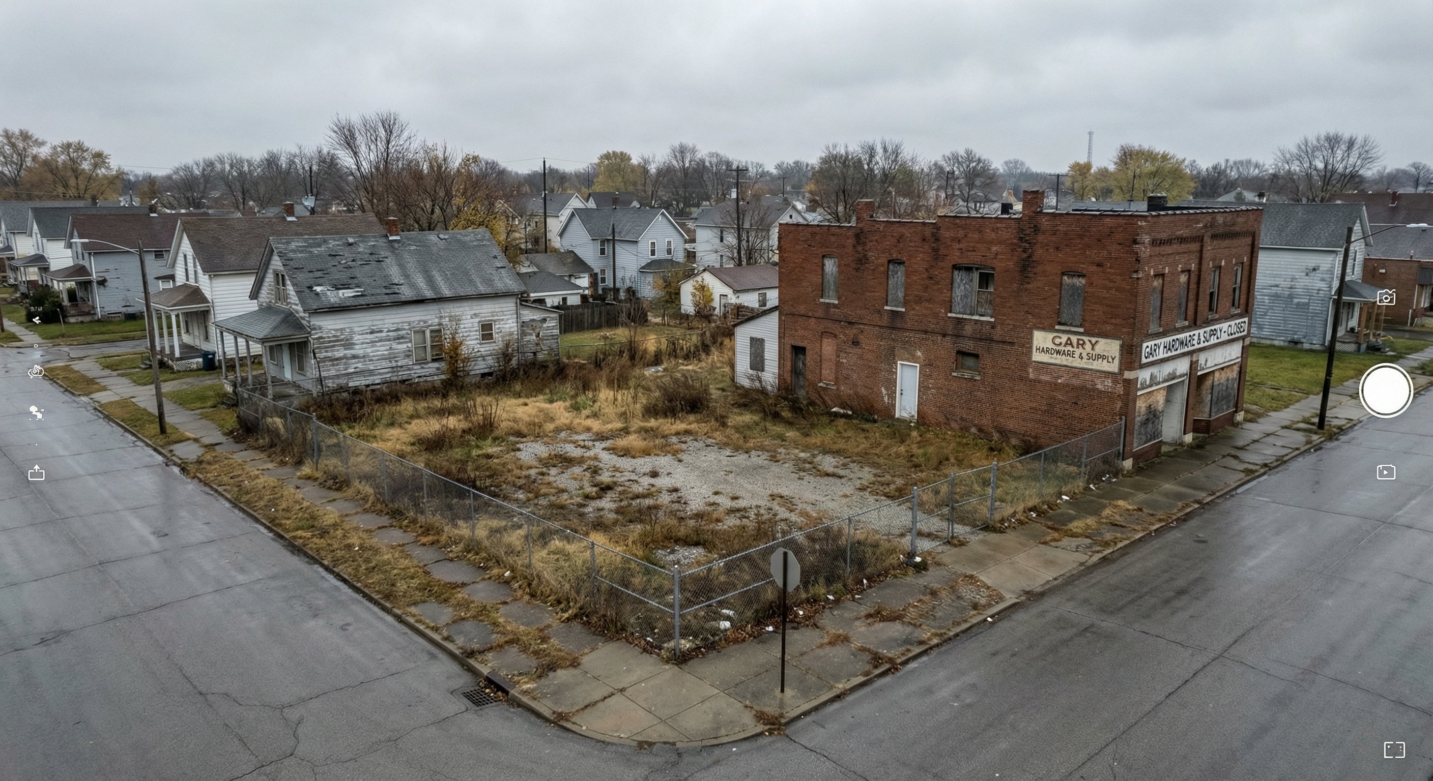 Aerial view of a vacant corner lot in a Rust Belt Indiana city with chain-link fence and abandoned commercial building nearby