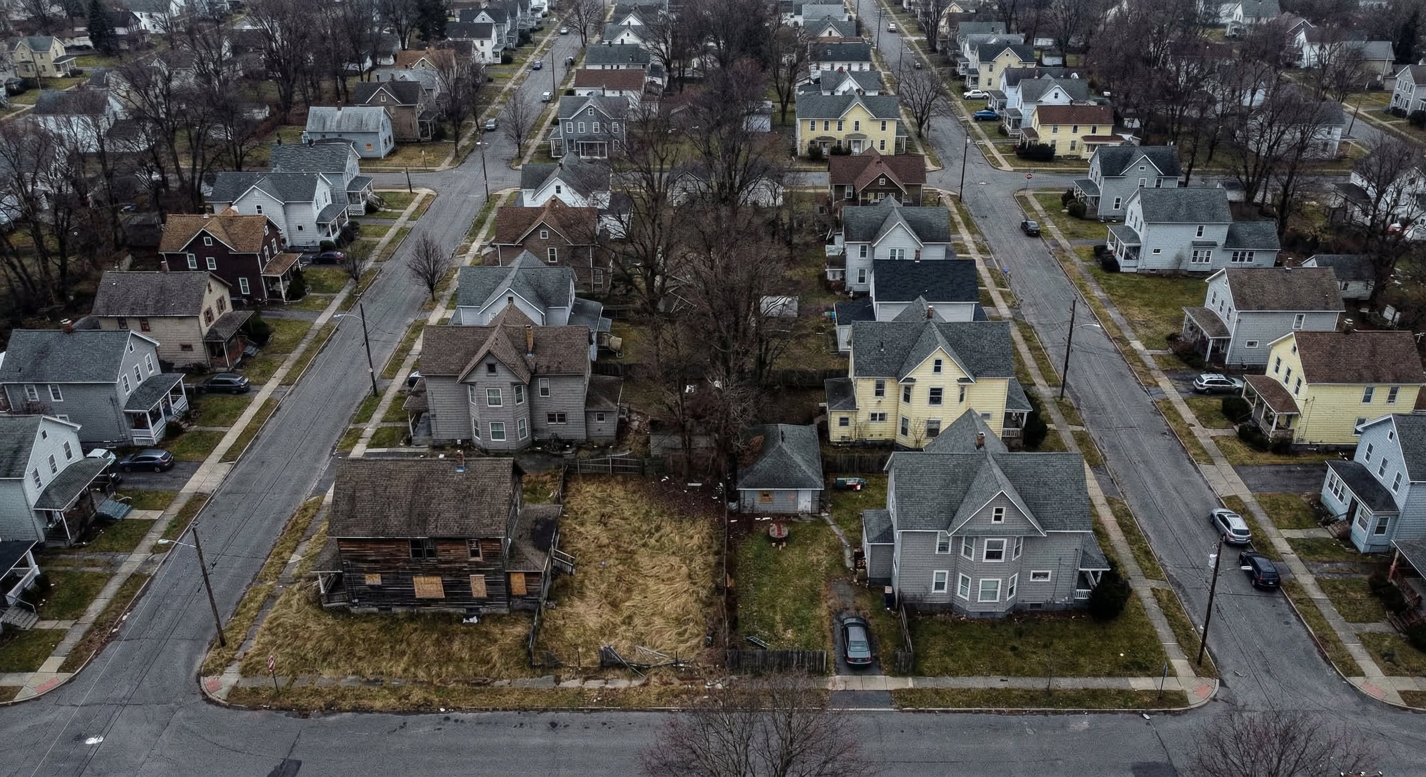 Aerial view of a residential neighborhood in upstate New York with rows of two-story wood-frame homes and bare maple trees lining narrow streets, several vacant properties with overgrown lots visible among occupied homes
