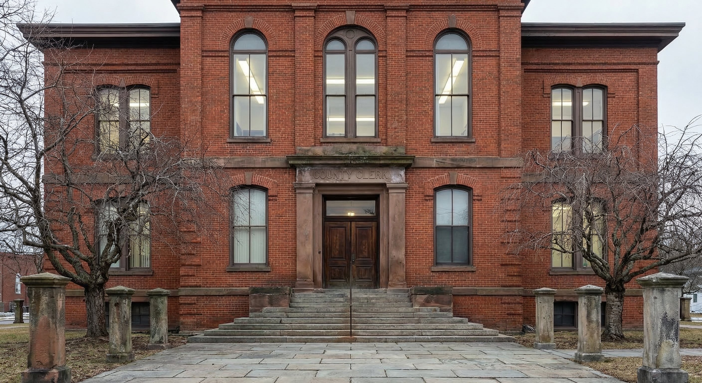 A wide-angle exterior photograph of a red-brick county courthouse in upstate New York with stone steps, tall double doors, and County Clerk signage above the entrance, bare winter trees flanking the building, gray overcast sky