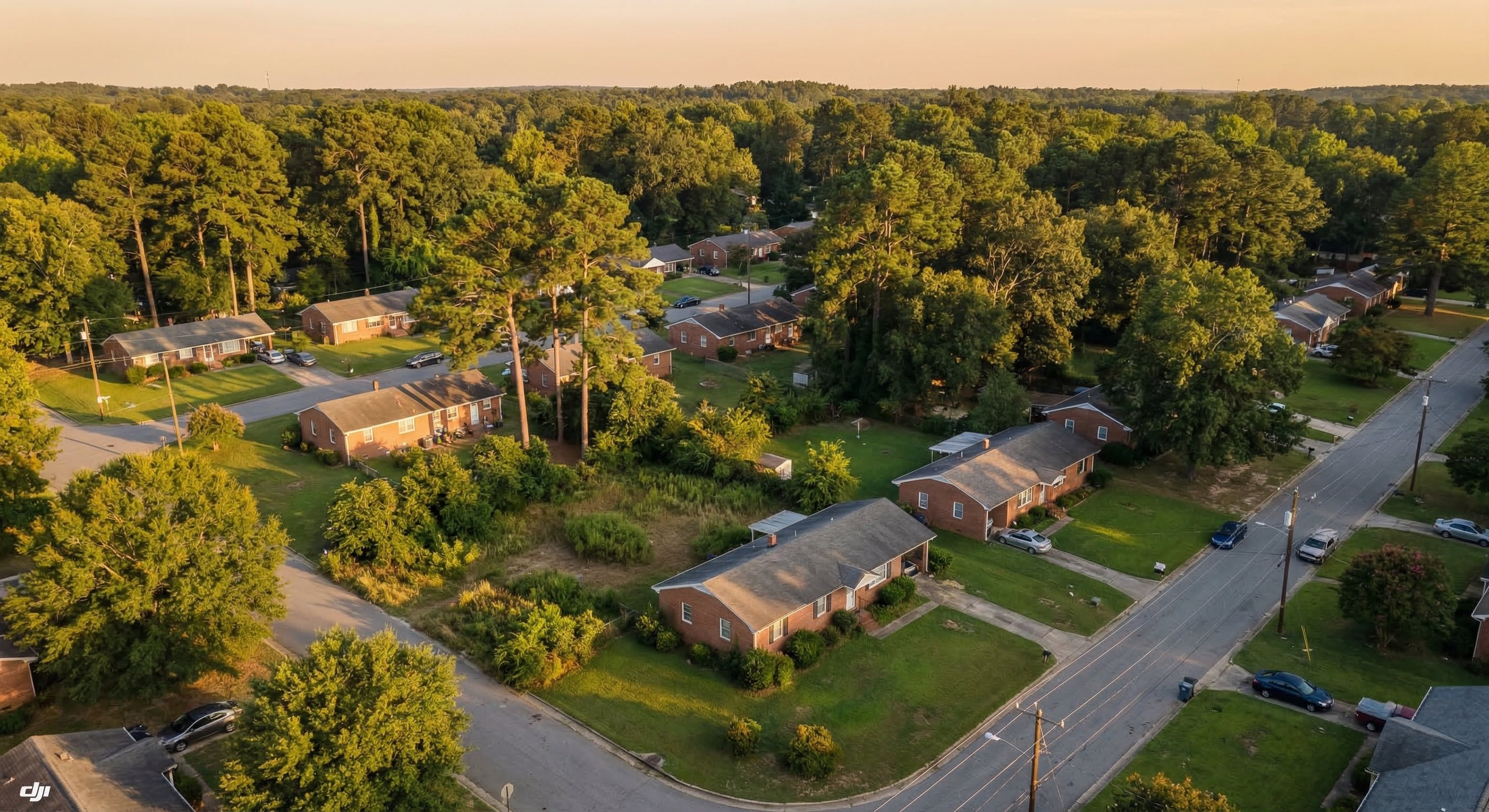 Aerial drone photograph of a mixed residential neighborhood in Durham, North Carolina, showing older brick ranch homes with mature pine and oak tree canopies, some with maintained yards and some with visible neglect, golden afternoon light